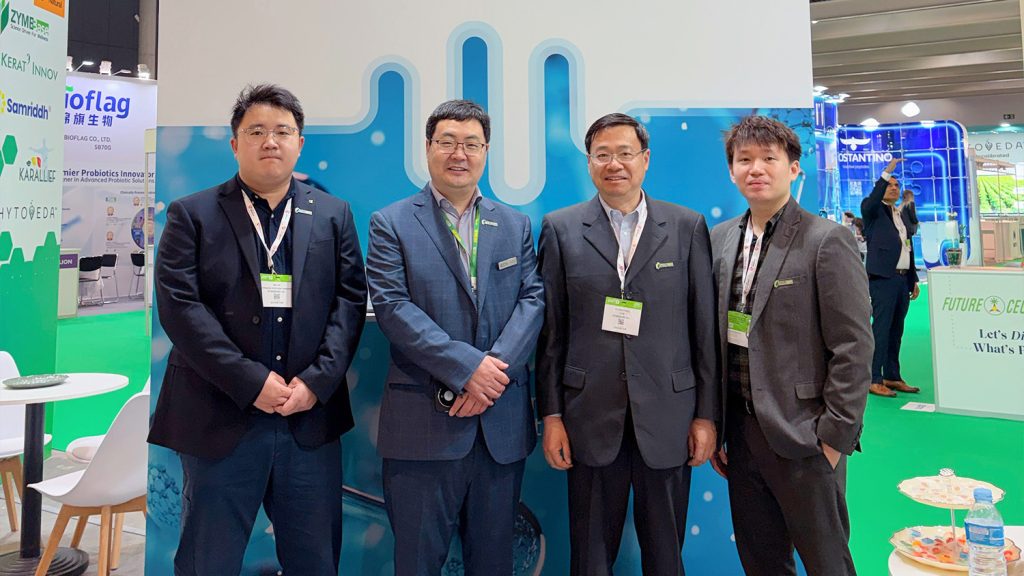 Four men in business suits posing at a blue exhibition booth at a trade show, wearing conference badges.